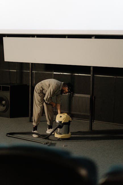 A professional cleaning technician dressed in full protective gear, including a white hazmat suit, mask, and gloves, performing deep cleaning of a beige carpet in a residential living room. The techni