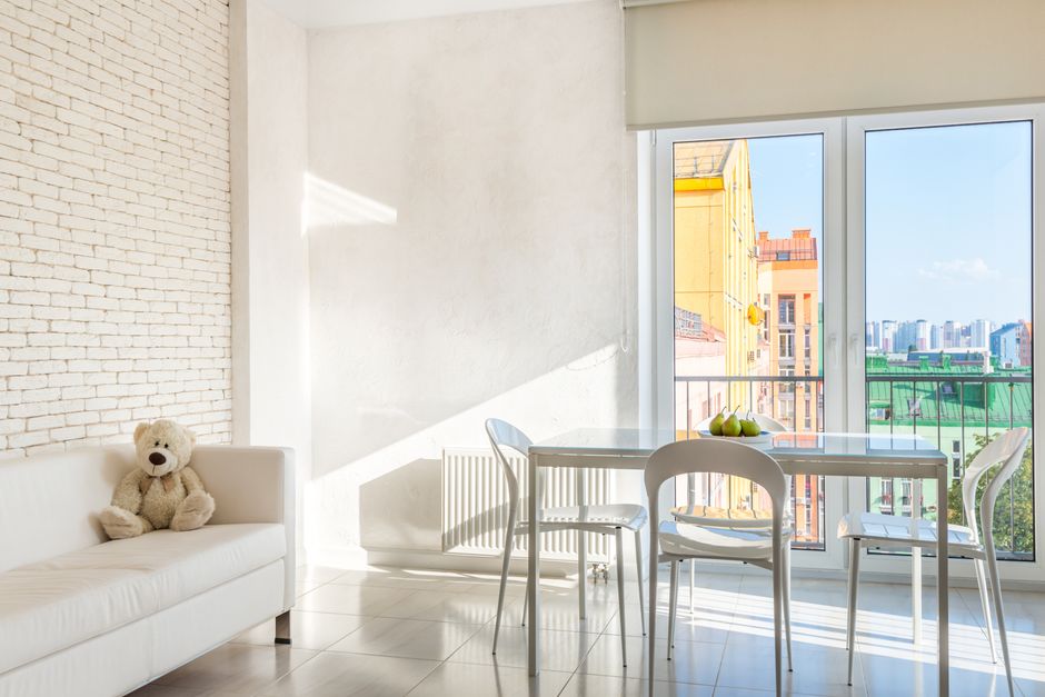 A bright, modern dining area with a white table and four matching white chairs positioned near large glass sliding doors that lead to a balcony. The table holds a decorative bowl of green apples. The 