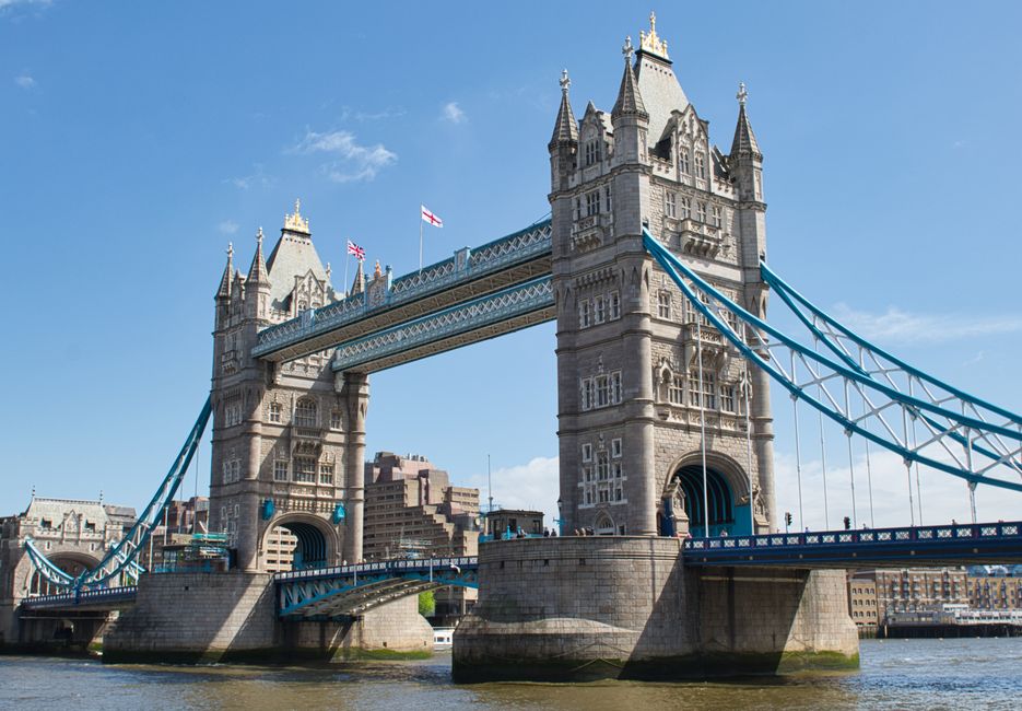 A detailed aerial view of Tower Bridge in Bermondsey, London, showcasing its iconic twin towers with Gothic Revival architecture, connected by a blue suspension span across the River Thames. The bridg